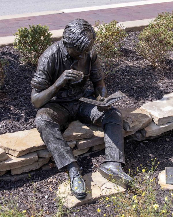 Statue of a kid reading a book while eating a burg...
